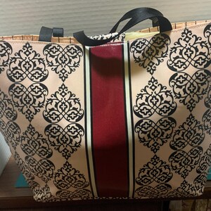 Może przedstawiać: Duża torba typu tote z beżowym tłem i czarnym wzorem ornamentalnym. Pionowy bordowy pas biegnie przez środek, obramowany cienkimi białymi liniami. Torba ma czarne uchwyty.