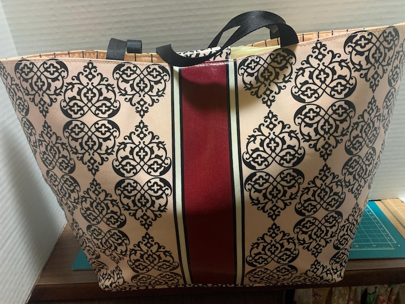 Może przedstawiać: Duża torba typu tote z beżowym tłem i czarnym wzorem ornamentalnym. Pionowy bordowy pas biegnie przez środek, obramowany cienkimi białymi liniami. Torba ma czarne uchwyty.