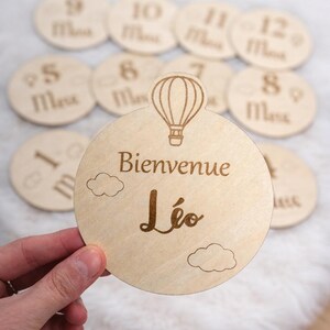 Può includere: Dischi di legno per le tappe del bambino. Il disco principale dice "Bienvenue Léo" con un'illustrazione di mongolfiera e disegni di nuvole. Dischi aggiuntivi numerati da 1 a 12 con il testo "Mois".