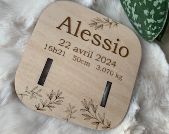 Puede incluir: Placa de madera clara grabada con el nombre "Alessio", la fecha "22 avril 2024", la hora, una longitud de 50 cm y un peso de 3,070 kg. Presenta diseños florales decorativos en las esquinas.