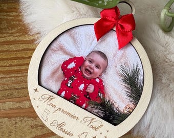 May include: A wooden Christmas ornament with a photo of a baby wearing a red onesie. The ornament has a red bow and the text "Mon premier Noel Alessio".