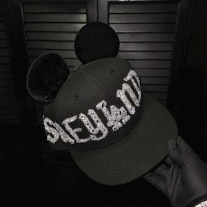 May include: Black baseball cap with the word "Disneyland" in a white, ornate font across the front. The cap has two black, plush mouse ears attached to the top. The brim is curved, and the cap is being held by a gloved hand.