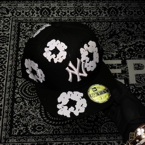 Op de afbeelding: Zwarte baseballcap met witte bloemmotieven en het witte New York Yankees-logo. De cap heeft een gebogen klep en een gele sticker met maat 7 1/4.