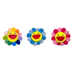 Op de afbeelding: Drie pluche bloemornamenten met een geel rond gezicht, een brede rode lachende mond en zwarte ogen. De linkerbloem heeft veelkleurige bloemblaadjes, de middelste bloem heeft blauwe bloemblaadjes en de rechterbloem heeft roze en rode bloemblaadjes.