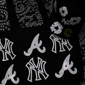 Op de afbeelding: Zwarte baseballcap met witte teamlogo's, waaronder de Atlanta Braves en New York Yankees. De cap heeft een 59Fifty-sticker en een bloemenontwerp aan de zijkant. De achtergrond heeft een zwart-wit paisley patroon.