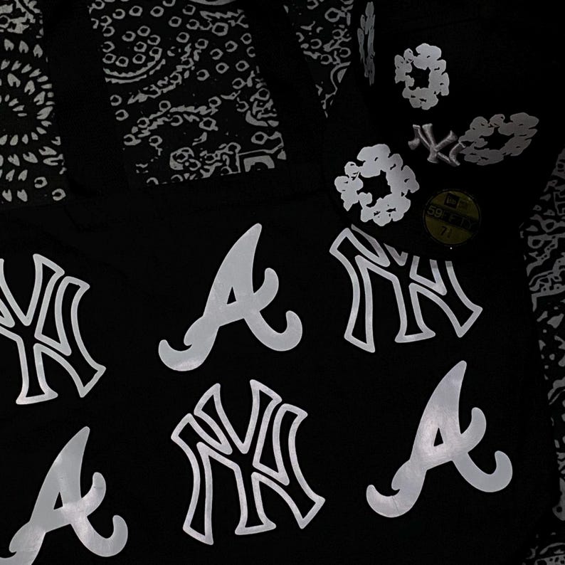 Op de afbeelding: Zwarte baseballcap met witte teamlogo's, waaronder de Atlanta Braves en New York Yankees. De cap heeft een 59Fifty-sticker en een bloemenontwerp aan de zijkant. De achtergrond heeft een zwart-wit paisley patroon.