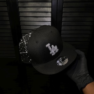 May include: A black snapback cap featuring a prominent silver-toned rhinestone-encrusted LA logo on the front crown. The side panel is decorated with a scattered pattern of clear rhinestones. A circular 9FIFTY sticker is affixed to the flat brim.