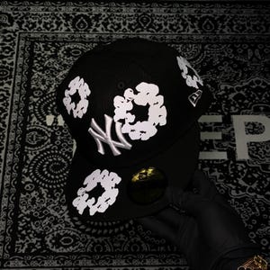 Op de afbeelding: Een zwarte baseballcap met witte bloempatronen en het zilveren New York Yankees-logo. De cap heeft een gebogen klep en een klein New Era-logo. De cap wordt vastgehouden door een gehandschoende hand.