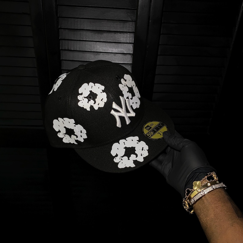 Op de afbeelding: Zwarte baseballcap met witte bloemmotieven en het logo van de New York Yankees. De cap heeft een gele sticker op de klep. De cap wordt vastgehouden door een gehandschoende hand.