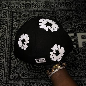 Op de afbeelding: Zwarte baseballcap met witte bloemmotieven. De cap heeft een gebogen klep en een kleine MLB-logopatch. De cap wordt getoond tegen een patroonachtergrond.