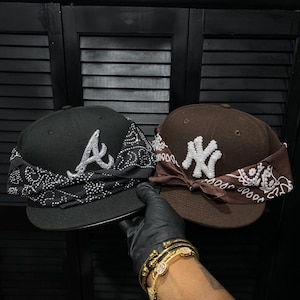 May include: Two baseball caps are shown, one black and one brown. The black cap has a white "A" and a black and white bandana. The brown cap displays a white "NY" logo and a brown and white bandana.