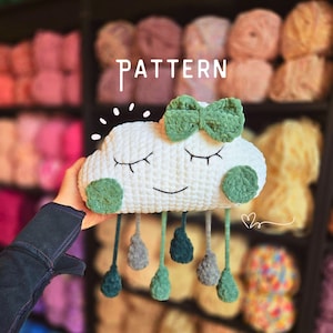 May include: A white crochet cloud with a green bow and accents, featuring a smiling face and closed eyes. The cloud has green circles on the sides and dangling teardrops in shades of green and grey. The word "PATTERN" is above the cloud.