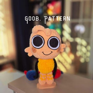 Könnte beinhalten: Eine plüschige, orangefarbene Puppe mit großen schwarzen und weißen Augen, kleinen Armen und einem lächelnden Gesicht. Die Puppe hat einen gelben Torso und gemusterte gelbe Beine. Der Text "goob. PATTERN" steht über der Puppe.