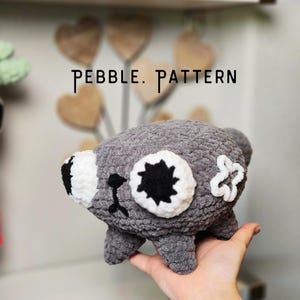 Op de afbeelding: Een grijs gehaakt knuffeltje met grote witte ogen met zwarte stervormige pupillen, een kleine zwarte neus en een gestikte mond. De tekst PEBBLE. PATTERN is zichtbaar boven het speelgoed, dat een getextureerd oppervlak van garen heeft.