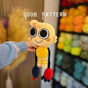 Puede incluir: Un peluche de ganchillo hecho a mano con cabeza de color amarillo pálido, grandes ojos blancos y negros, una pequeña sonrisa y manchas marrones en las mejillas. El cuerpo es amarillo con brazos negros, un pie azul oscuro y otro rojo. Texto: GOOB. PATTERN.