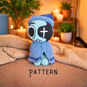May include: A plush doll made of soft blue and light green yarn, featuring a large round head with one circular eye and one eye marked with a white cross. The doll wears a blue hooded cape and has the word PATTERN printed below it in black text.