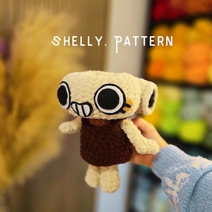 Op de afbeelding: Een gehaakt knuffeltje met een crèmekleurig hoofd, grote zwarte ronde ogen en een kleine mond, gekleed in een donkerbruine jurk. De tekst SHELLY. PATTERN is erboven zichtbaar. Het speelgoed heeft kleine armen en benen.