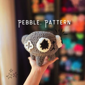 Op de afbeelding: Een gehaakte grijze koala beer met witte en zwarte accenten, vastgehouden in een hand. De koala heeft een lachend gezicht en een bloemachtig ontwerp aan de zijkant. De tekst "PEBBLE. PATTERN" staat boven de koala.