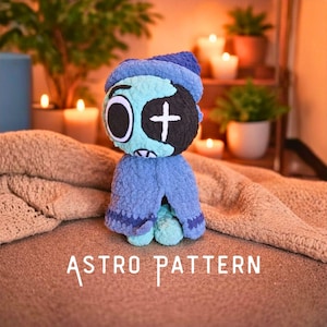 Op de afbeelding: Handgemaakte blauwe en turquoise gehaakte pop met een zwart ooglapje en een wit kruis. De pop heeft een blauwe hoed en cape. De tekst "ASTRO PATTERN" staat in witte letters onderaan de afbeelding. De pop zit op een bruin oppervlak.