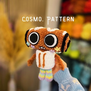 Pode incluir: Um brinquedo de pelúcia de crochê marrom com grandes olhos pretos e brancos, um moletom branco e pernas multicoloridas. O texto COSMO. PATTERN é visível acima do brinquedo. A pelúcia apresenta pequenos detalhes florais nas laterais.