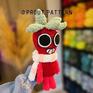 May include: A red crocheted plush toy shaped like a strawberry with light green leaves on top, large black circular eyes, and a white and pink striped scarf. The text SPROUT.PATTERN is visible above the plush toy in black lettering.