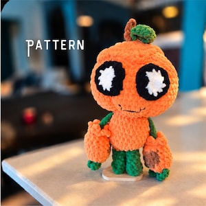 Puede incluir: Una figura de calabaza de ganchillo con una cabeza naranja grande, ojos negros y blancos y una boca pequeña. Tiene piernas verdes, brazos con manos naranjas y un pequeño tallo verde en la parte superior. La palabra PATTERN aparece en blanco.