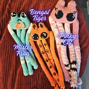 May include: Three handmade crochet plush toys: a teal frog with a bow tie, an orange Bengal tiger, and a pink cat. Each toy has large, expressive eyes and long, dangling limbs. The text on the image reads "Mister Frog", "Bengal Tiger", and "Pinky Cat".