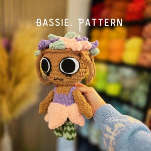 May include: A brown crocheted doll with large black eyes and a small mouth, wearing a purple top and a light pink skirt. The doll has a crown of pastel green, purple, and pink yarn loops on its head. The text BASSIE. PATTERN is visible above.