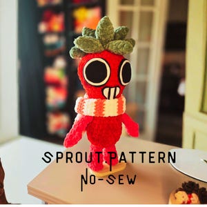 Può includere: Bambola rossa fatta a mano all'uncinetto con una testa verde a forma di foglia, grandi occhi neri e una sciarpa a righe. Il testo "SPROUT PATTERN NO-SEW" è in nero. Si trova su una piccola base di legno.