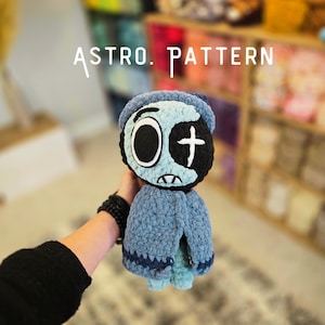 May include: A handmade crochet plush doll featuring a light blue and black color scheme with a beanie hat. The doll has one large circular eye and one eye with a white cross symbol. The text ASTRO. PATTERN is visible above the plush doll.