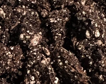 May include: Close-up of potting soil mix, featuring dark brown organic matter with visible white perlite particles. The texture appears coarse and well-draining, suitable for various plant types. The mix includes small pieces of organic material.