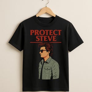 以下が含まれることがあります： 黒いTシャツ。赤い文字で「PROTECT STEVE」と書かれ、サングラスと緑色のジャケットを着た男性のイラストが描かれています。木製のハンガーに掛けられています。