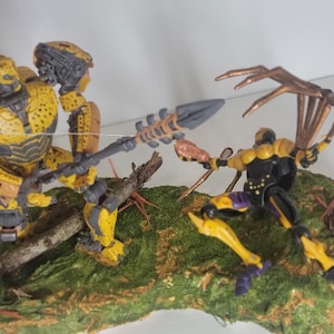 以下が含まれることがあります： 黄色と黒のロボットのアクションフィギュアが、長い尻尾と複数の脚を持つ黄色と黒のクモのようなアクションフィギュアと戦っています。両方のフィギュアは、緑と茶色のベースにダイナミックな戦闘姿勢で配置されています。