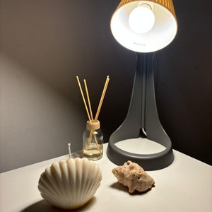 May include: A white seashell-shaped candle sits on a white surface next to a small glass bottle with wooden sticks and a brown seashell. A gray lamp with a brown pleated shade is in the background.