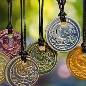 May include: A collection of ceramic pendant necklaces with a celestial design. Each pendant features a moon and cloud motif in various colors, including blue, green, and gold, strung on black cords. The pendants are round with a hole at the top.