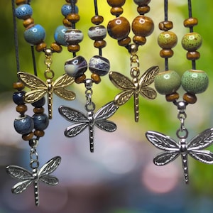 May include: Close-up of several beaded necklaces with dragonfly pendants. The necklaces feature a variety of beads in shades of blue, brown, and green. The dragonfly pendants are silver and gold-toned, with intricate wing details. The background is a soft, blurred green.