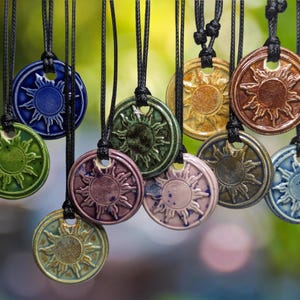 Pode incluir: Uma coleção de dez pingentes de cerâmica, cada um com um design de sol estilizado em várias cores, incluindo azul, verde, marrom, roxo e dourado. Os pingentes são encordoados em cordões pretos.