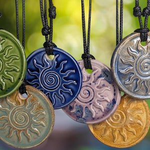 Può includere: Una collezione di collane con ciondoli a forma di sole in ceramica in vari colori, tra cui verde, blu e oro. Ogni ciondolo presenta un disegno a forma di sole con un centro a spirale ed è appeso a un cordino nero.
