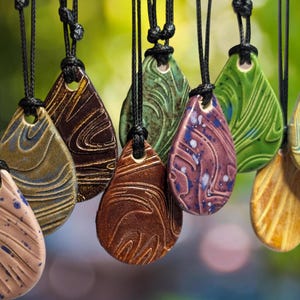 May include: A collection of teardrop-shaped ceramic pendants in various colors, including brown, green, and purple, each with a unique swirled pattern. The pendants are suspended from black cords, creating a display of handcrafted jewelry.