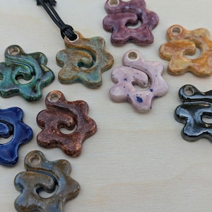 May include: A selection of ceramic pendants in shades of blue, green, brown, and purple. Each pendant features an organic design with a hole for threading. One pendant is attached to a black cord.
