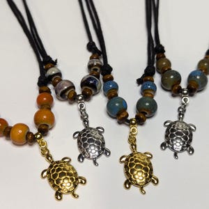 May include: Three necklaces with a turtle pendant. Each necklace has brown wooden beads and colorful ceramic beads. The turtles are silver, gold, and silver.