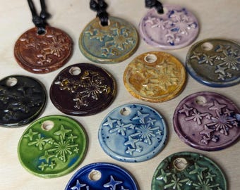 May include: A collection of 11 ceramic pendants featuring a snowflake design. The pendants come in a variety of colours, including green, blue, purple, brown, and black. Each pendant has a hole at the top for hanging.