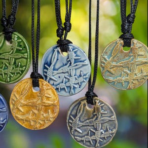 May include: A collection of ceramic pendant necklaces with a barbed wire design. The pendants are circular and come in various colors, including green, blue, and gold. Each pendant hangs from a black cord.