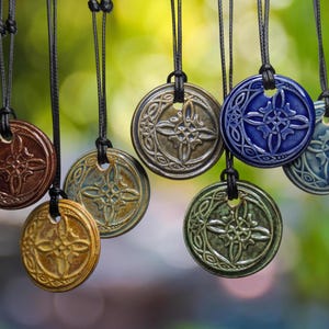 May include: A collection of ceramic pendant necklaces in various colors, including brown, gold, blue, and green. Each pendant is round with a raised, symmetrical design and hangs from a black cord. The background is a soft, blurred green.