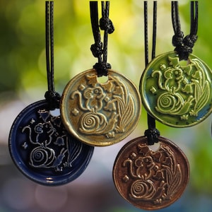 May include: Close-up of several ceramic pendant necklaces. Each pendant features a circular design with a relief image of a monkey, flowers, and a spiral. The pendants are in various colors, including blue, green, brown, and gold, and are suspended from black cords.