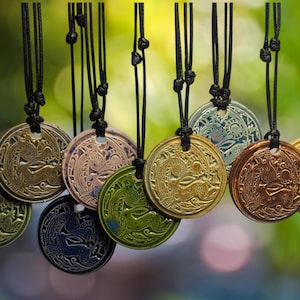 以下が含まれることがあります： 精巧なエンボス加工が施された、丸いペンダントネックレスのコレクション。ペンダントは、金、ブロンズ、銀など、さまざまなメタリックカラーで、黒いコードで吊り下げられています。背景は柔らかくぼやけた緑色です。