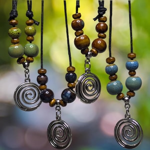 以下が含まれることがあります： 茶色、青、緑色のビーズチェーンと、渦巻きペンダントのネックレス。銀色のペンダントは、さまざまなサイズと質感のビーズと組み合わされています。ネックレスは、ぼやけた緑色の背景に配置されています。