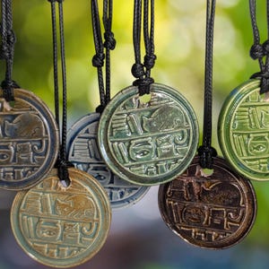 May include: A collection of ceramic pendant necklaces in various colors, including blue, green, and brown. Each pendant features a circular design with an embossed cat and hieroglyphic-style symbols, suspended from black cords. The background is a soft, blurred green.