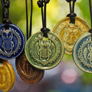 Puede incluir: Una colección de collares con colgantes de cerámica en varios colores, incluyendo azul, verde y dorado. Cada colgante presenta un diseño detallado de escarabajo y está ensartado en un cordón negro. Los colgantes son redondos con un borde decorativo.