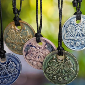 May include: Several ceramic pendant necklaces featuring a moth and floral design. The pendants are in shades of blue, green, and pink, suspended from black cords. The background is a blurred green.